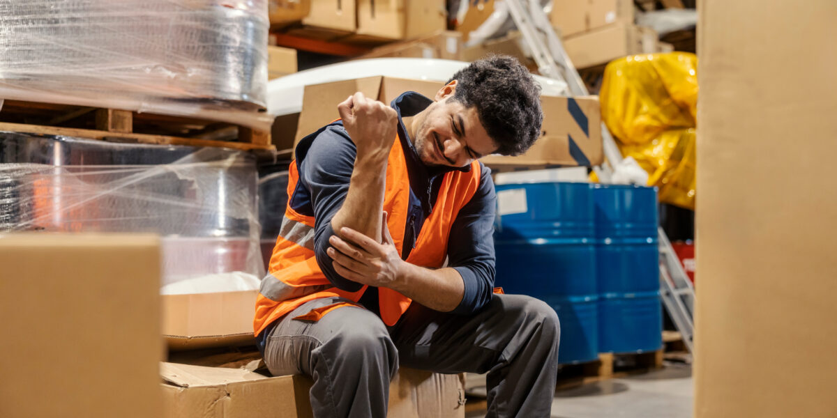 Warehouse worker with injured elbow sits amid boxes, illustrating need for work injury claims support in Queensland.