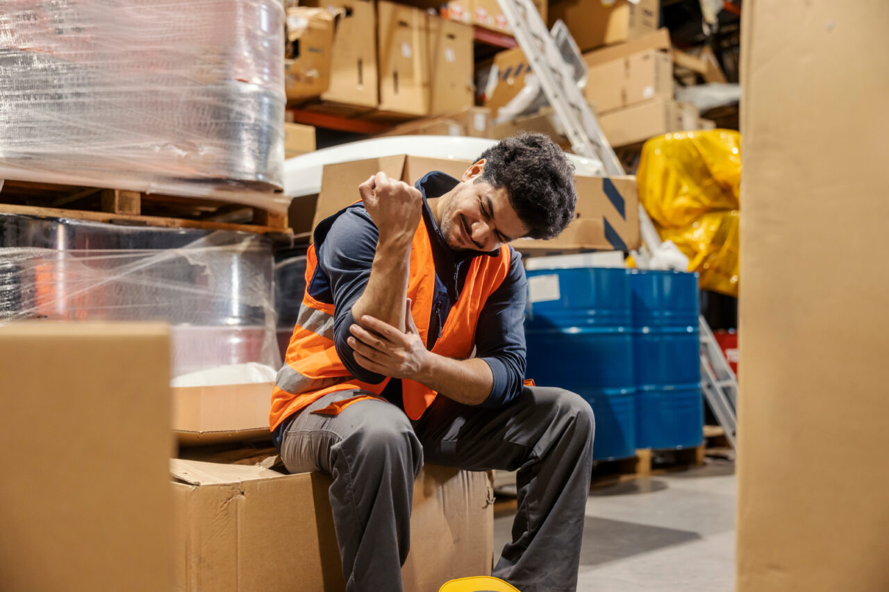 Warehouse worker with injured elbow sits amid boxes, illustrating need for work injury claims support in Queensland.